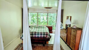 Un dormitorio con una cama, un aparador de madera, un ventilador y una ventana con vistas a la vegetación.
