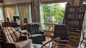 Una sala de estar acogedora con un sofá marrón, un sillón negro, una mecedora de madera, una estantería y una ventana con cortinas.