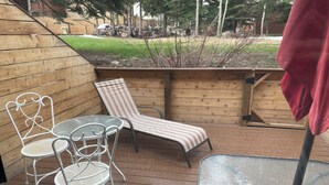 Un patio coperto con un tavolo e due sedie, una poltrona reclinabile a righe e vista su un giardino curato con alberi e un muro in pietra.