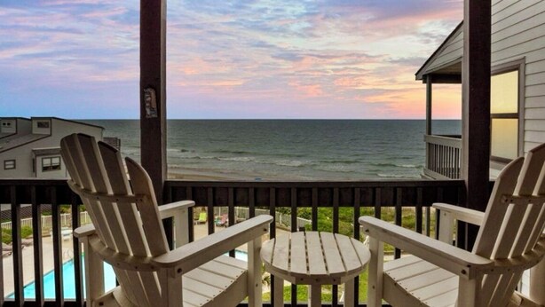A balcony with two Adirondack chairs and a small table, overlooking a beach and ocean during sunset.