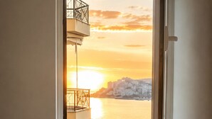 Un dormitorio con una cama, una mesita de noche y una ventana que ofrece vistas a un atardecer sobre el agua y edificios.
