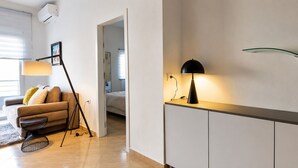 Una sala moderna con un sofá, una mesita auxiliar y una lámpara. Se ve un acceso a otra habitación.