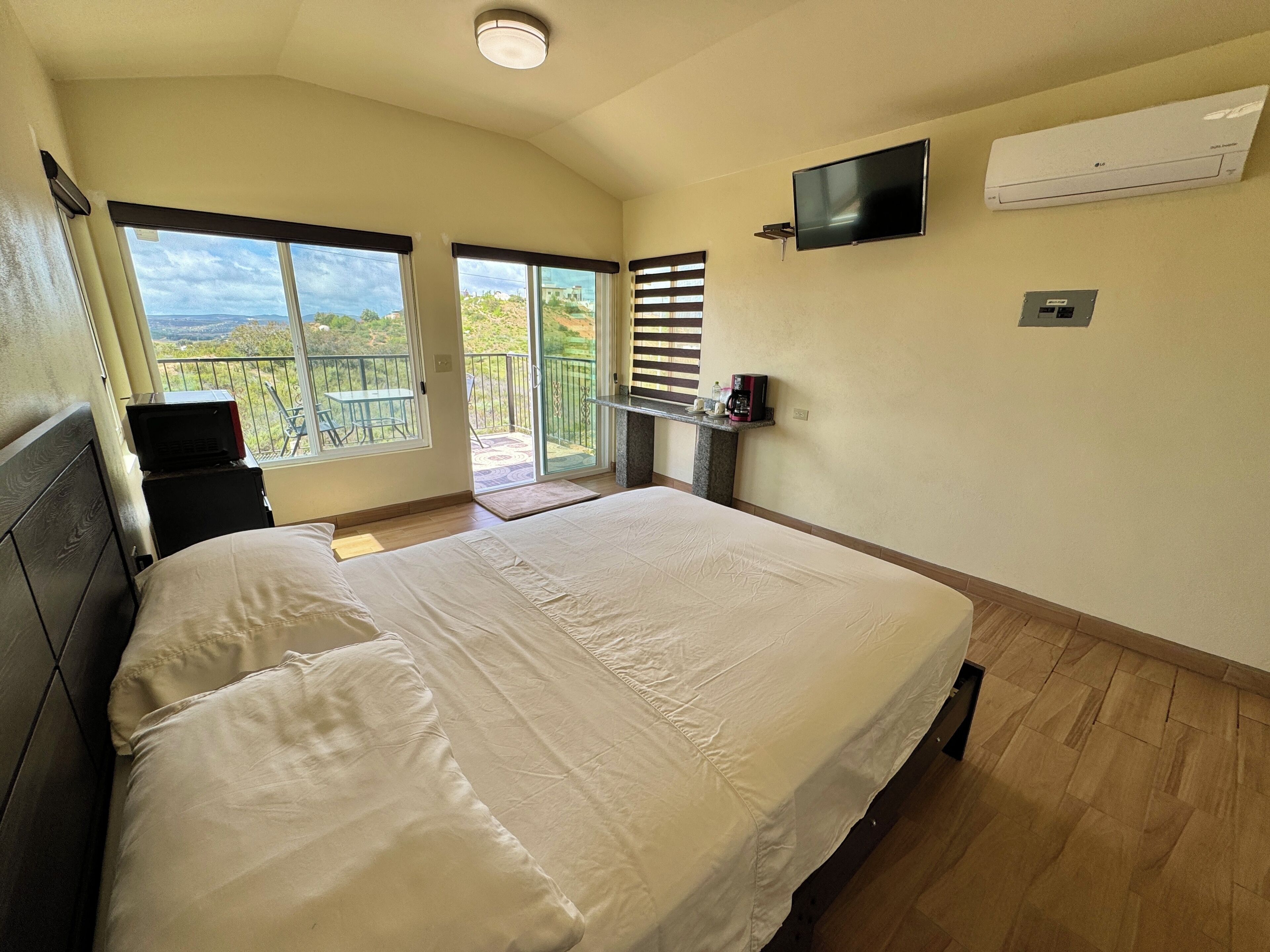 A bedroom with a large bed, a TV mounted on the wall, and a balcony with a view of the landscape.