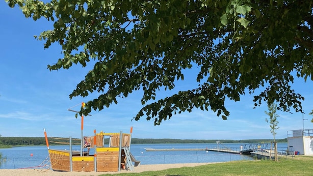 A wooden play structure by a lake with a dock and a small building in the background. - Holiday Home Reed Path (Warin)