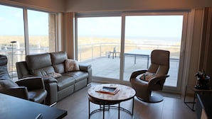 Een ruime woonkamer met een leren bank, een ronde salontafel en een fauteuil met hoge rugleuning. Grote ramen bieden uitzicht op het strand.
