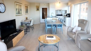 Een moderne woonkamer met een televisie, bank en salontafel. Er is een eethoek met stoelen en een tafel. De keuken is op de achtergrond zichtbaar.