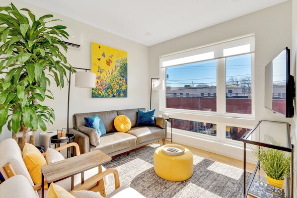 Una sala de estar moderna con sofá, reposapiés y sillones, un ventanal con vistas, un cuadro y una planta en maceta.