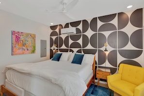 Ein Schlafzimmer mit einem Bett, einem Deckenventilator, einer wandmontierten Klimaanlage, einem Nachttisch mit Lampe und einem gelben Sessel.
