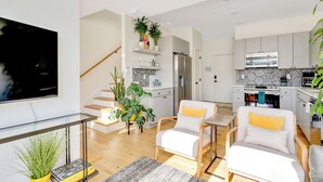 Een moderne woonkamer met een flatscreen televisie, twee witte banken met gele kussens, een salontafel, en een trap die naar een keuken leidt.