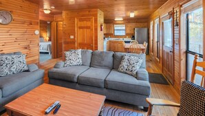 L'intérieur d'un chalet accueillant, avec un canapé gris, des murs en bois, un ventilateur de plafond et un coin cuisine visible à travers une porte ouverte.