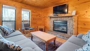 L’intérieur chaleureux d’un chalet avec une cheminée en pierre, des murs en bois, un téléviseur fixé au-dessus de la cheminée et un coin salon confortable avec un canapé et un fauteuil à bascule.