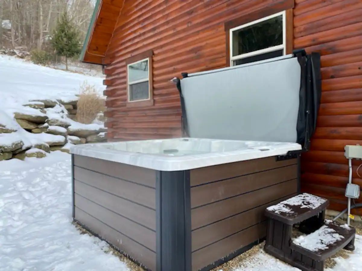 A hot tub in front of a wooden cabin with a stone wall and snow on the ground.