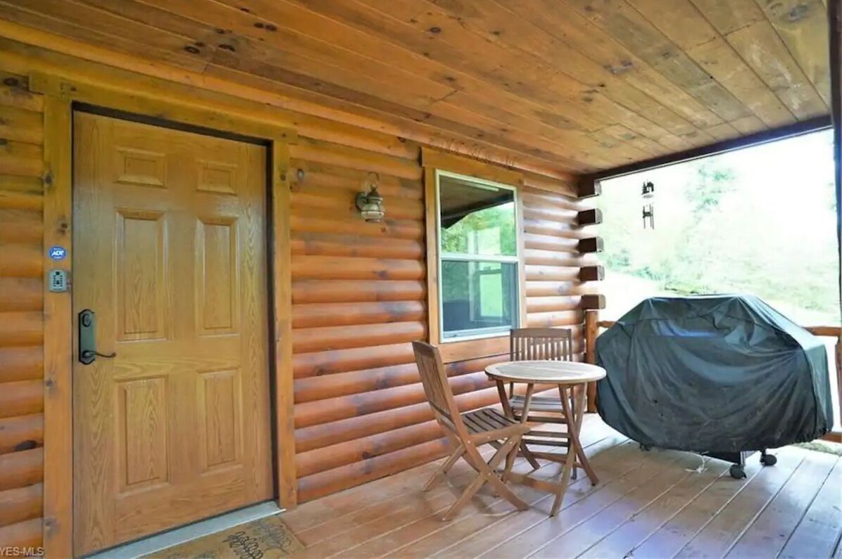 Une cabane en bois avec une grande porte, une fenêtre, un barbecue couvert et une petite table avec des chaises.