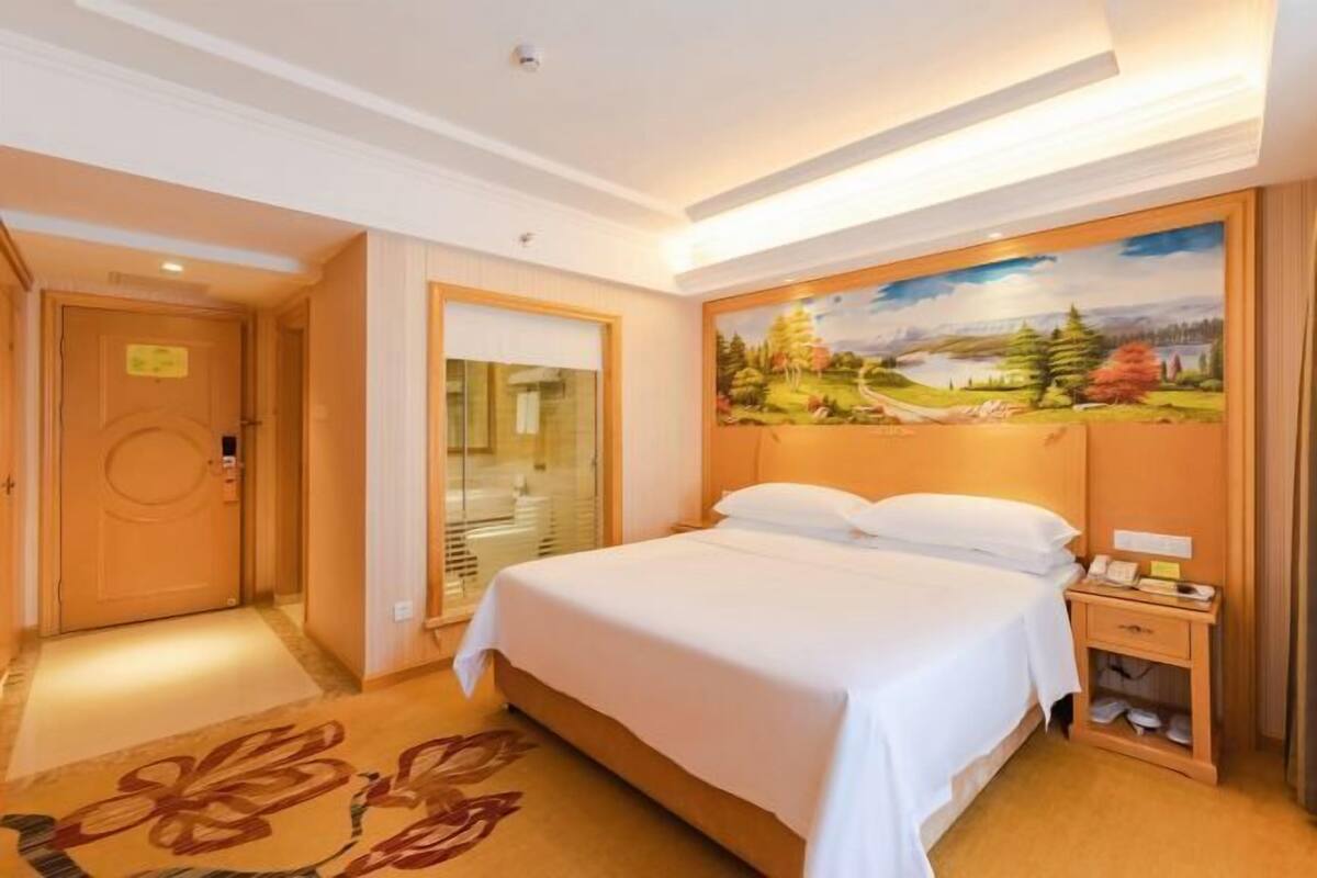 A hotel room with a bed, a painting on the wall, a nightstand, and a door.