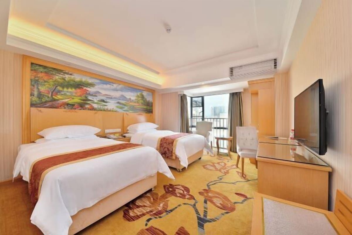 A hotel room with two beds, a flat-screen TV, a painting on the wall, and a view of the city.