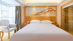 A hotel room with a large bed, a nightstand, a chair, and a large window with curtains. - Vienna Hotel (Shenzhen Gushu Metro Station) (Shenzhen)