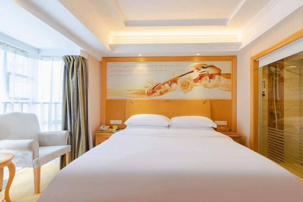 A hotel room with a large bed, a nightstand, a chair, and a large window with curtains.