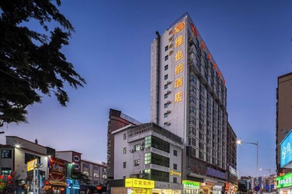 A multi-story hotel with a prominent vertical sign, surrounded by other buildings and illuminated signs. - Vienna Hotel (Shenzhen Gushu Metro Station) (Shenzhen)