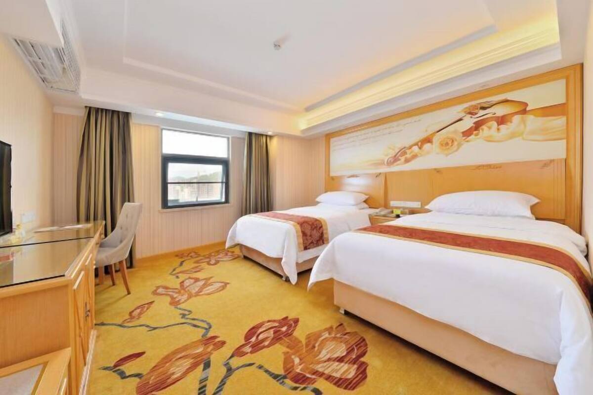 A hotel room with two beds, a desk with a chair, a large window, and a floral carpet.