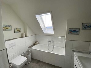 A small, white bathroom with a skylight, a bathtub, a toilet, and a trash can. - Holiday Home Ankerherz (Butjadingen)