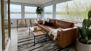 A spacious living room with a large window, a ceiling fan, a leather sofa, a coffee table, and a cactus plant. - Beautiful Private Lake Retreat - One of a kind! (Kawartha Lakes)