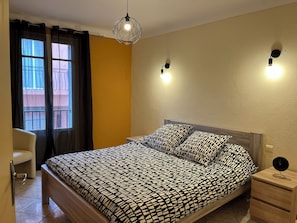 A bedroom with a bed, a chair, a bedside table with a clock, and a hanging light fixture. - Appartement Proche de la Mer, Idéal Pour 2 à 4 Personnes, Garage Privé (Collioure)