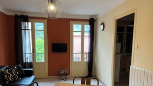 A room with a wooden dining table, a black sofa with patterned cushions, a TV mounted on the wall, and large windows with curtains. - Appartement Proche de la Mer, Idéal Pour 2 à 4 Personnes, Garage Privé (Collioure)