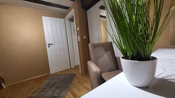 A room with a wooden floor, a grey rug, a white door, a beige armchair, and a white table with a potted plant.