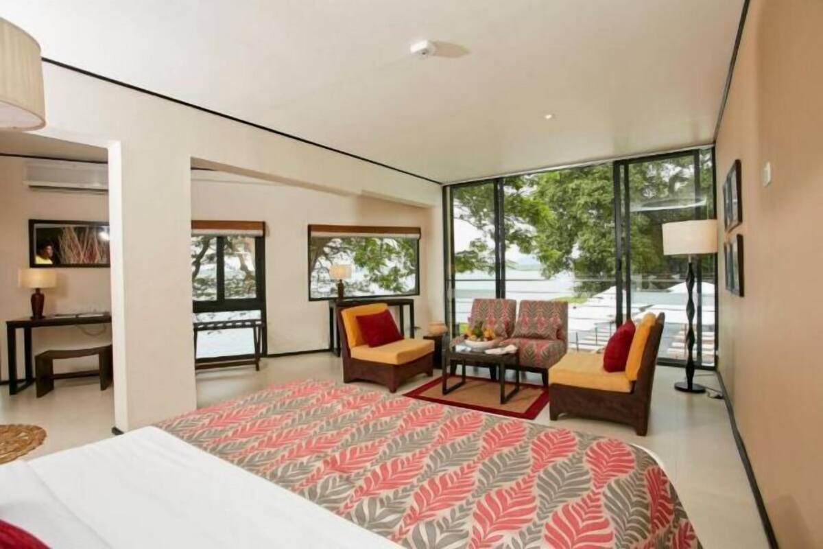 Une chambre d’hôtel moderne, dotée d’un grand lit, de deux fauteuils, d’une petite table et d’une grande fenêtre donnant sur l’extérieur.
