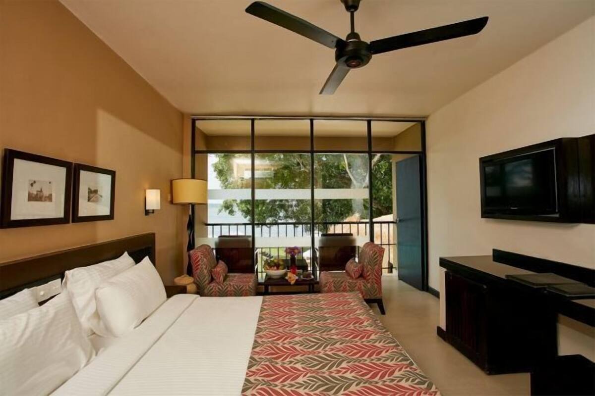 Une chambre d’hôtel avec un lit, un ventilateur de plafond, une télévision, un balcon avec des chaises et des tableaux encadrés au mur.