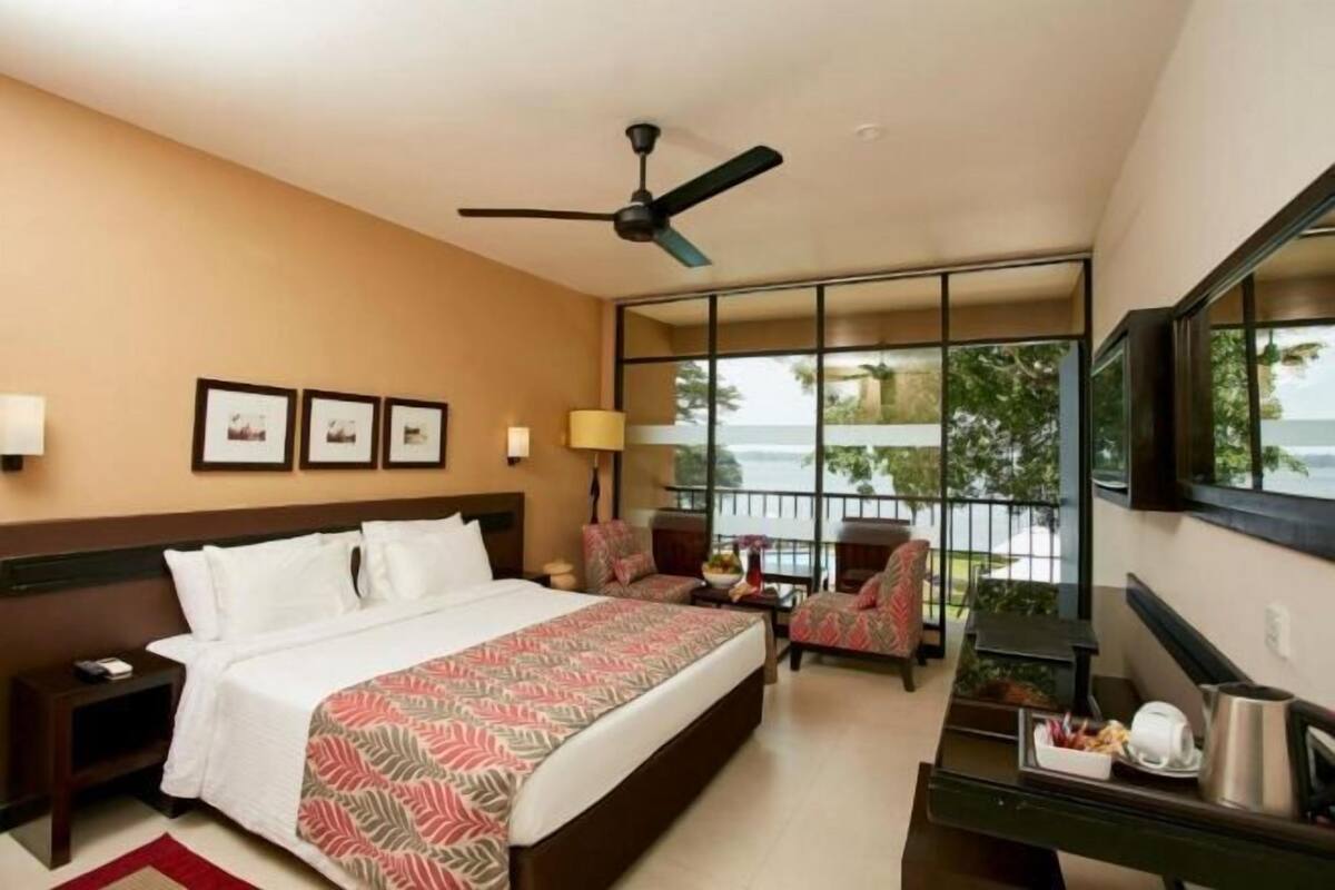 Une chambre d’hôtel équipée d’un lit, d’un bureau, d’une chaise, d’un ventilateur de plafond et d’une grande fenêtre donnant sur la mer.