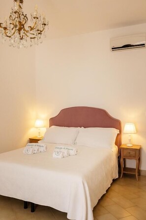 A neatly made bed with white linens and a pink headboard, two bedside lamps, and a chandelier.