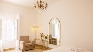 Une chambre d’hôtel avec un grand lit, un coin salon comprenant un fauteuil et une petite table, un miroir et un lustre.