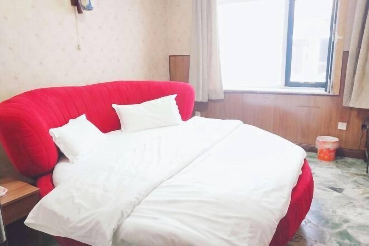 A room with a red bed, white bedding, a wooden nightstand, a window with curtains, and a marble floor.