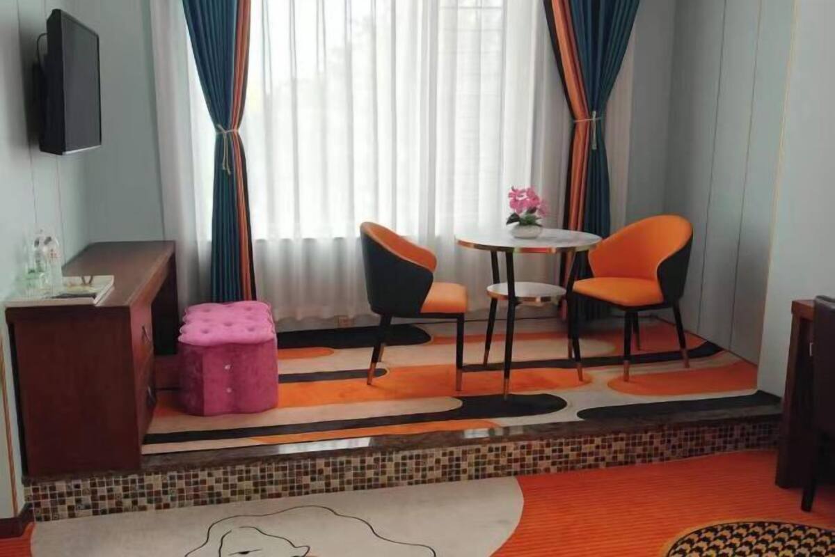 A hotel room with a small table and two chairs, a pink ottoman, a TV mounted on the wall, and a window with curtains.