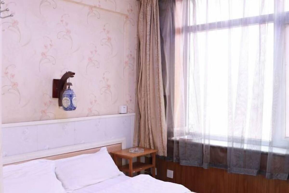 A bedroom with a bed, a small wooden table, a wall-mounted light fixture, and a window with curtains.
