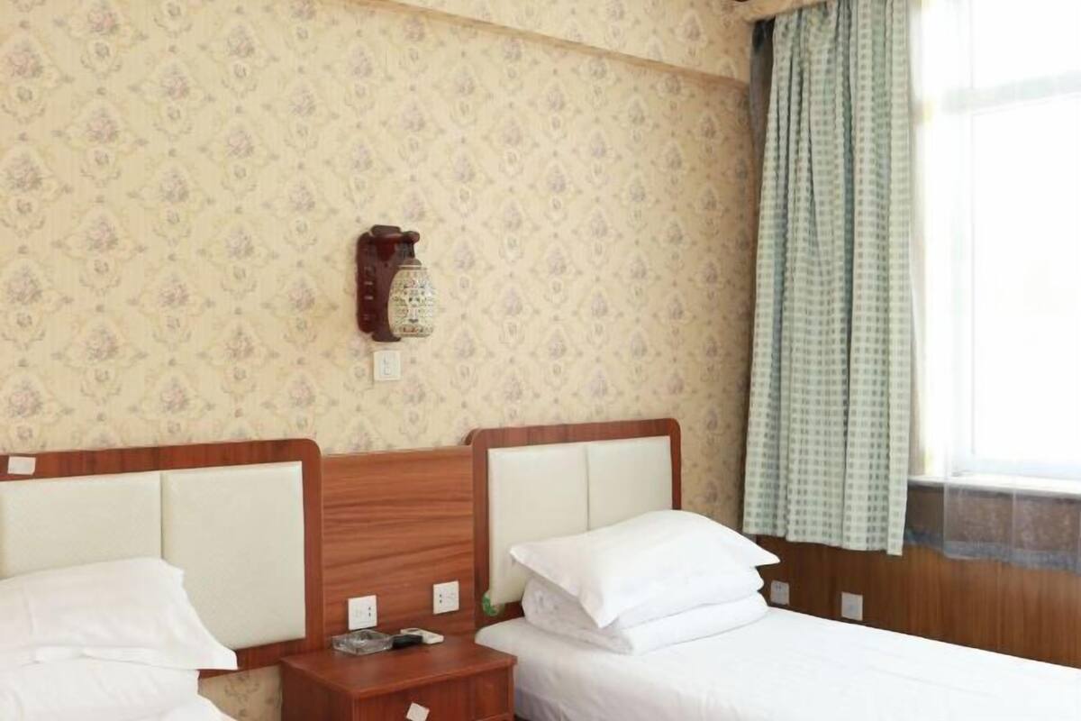 A hotel room with two beds, a nightstand, a wall-mounted lamp, and patterned wallpaper.