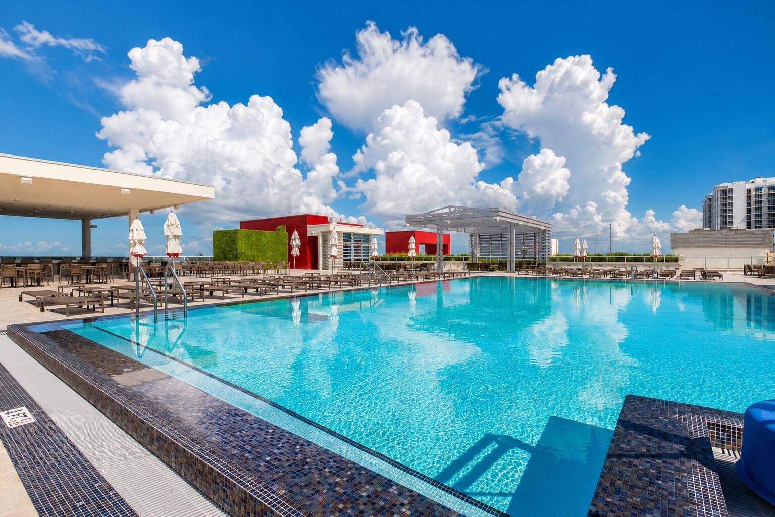 A swimming pool with lounge chairs and umbrellas, surrounded by buildings with a red and green facade.