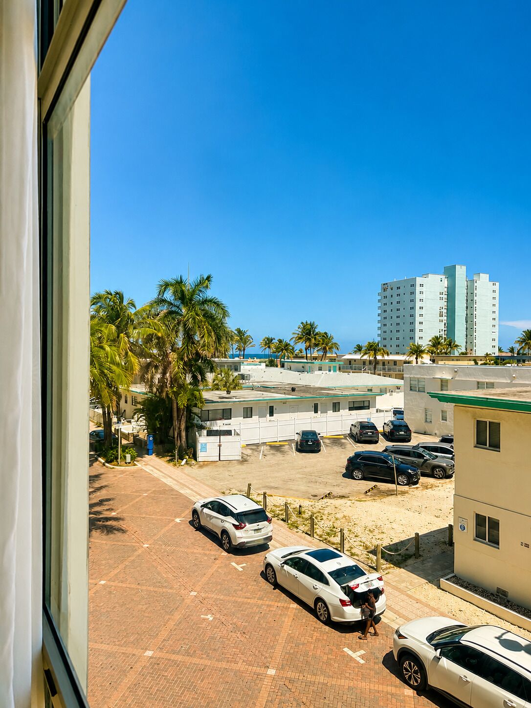 A parking lot with several cars, a building with a green roof, and palm trees in the background.