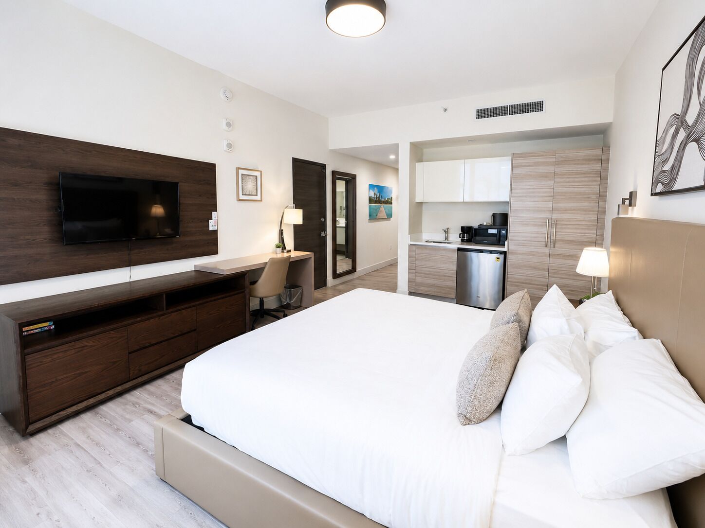 A modern hotel room with a large bed, a desk, a flat-screen TV, a kitchenette, and a bathroom.