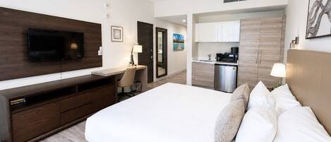 A modern hotel room with a large bed, a desk, a flat-screen TV, a kitchenette, and a bathroom.