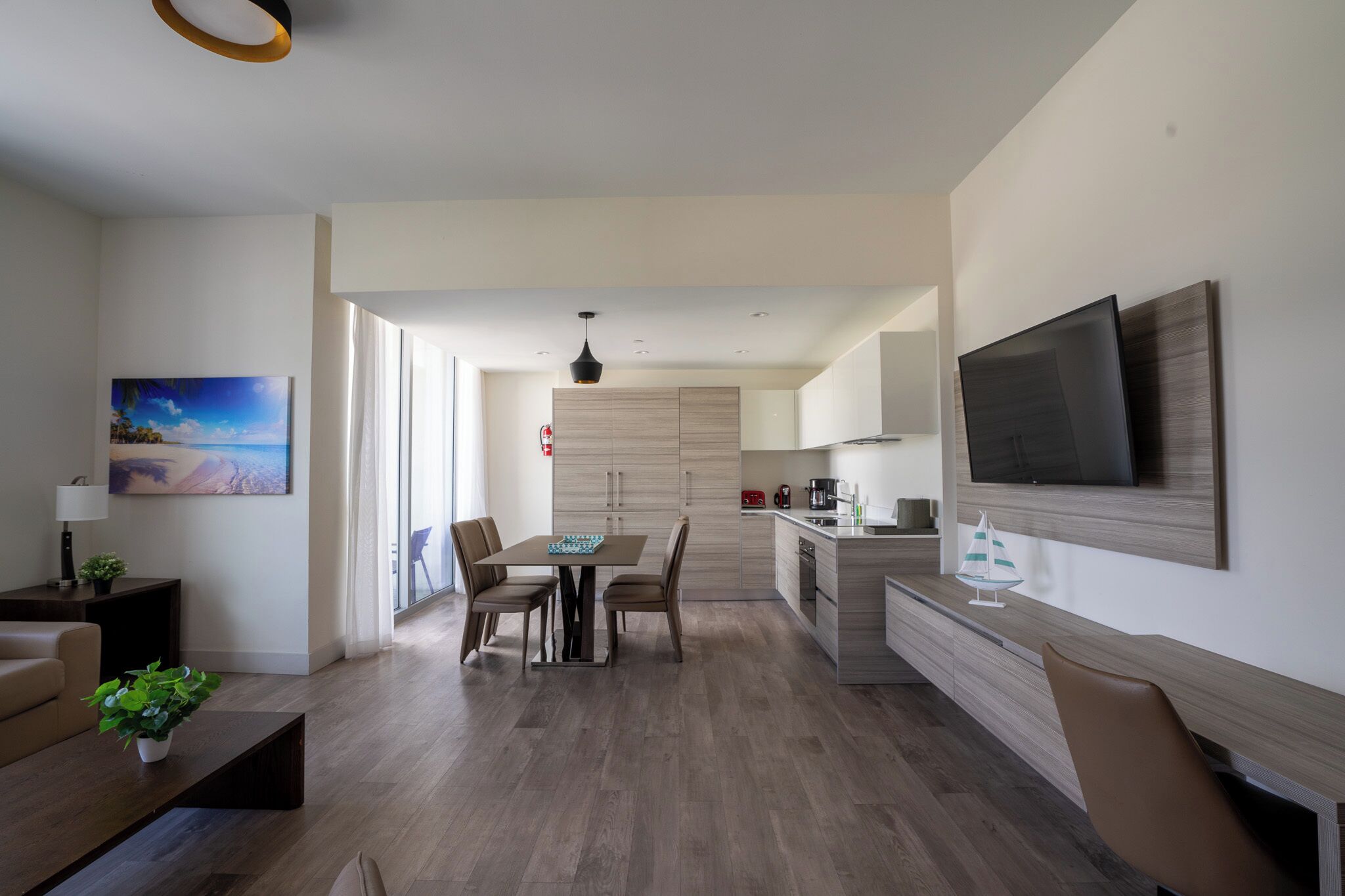 Ein modernes Wohnzimmer mit einer Couch, einem Couchtisch und einem an der Wand montierten Fernseher. Es gibt einen Essbereich mit Tisch und Stühlen. Die Küche ist sichtbar, inklusive Fernseher und einer Kaffeemaschine.