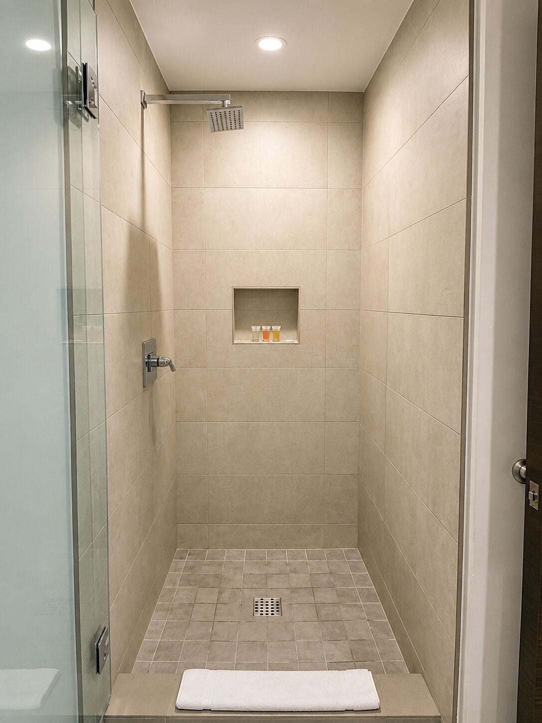 Ducha a ras de suelo con puerta de cristal, cabezal de ducha, estante con artículos de aseo y barra para toallas.