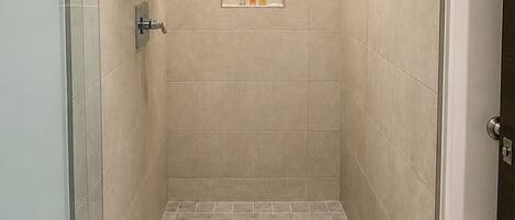 Ducha a ras de suelo con puerta de cristal, cabezal de ducha, estante con artículos de aseo y barra para toallas.