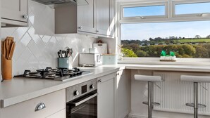 Une cuisine moderne avec des meubles blancs, une cuisinière à gaz, un four et une fenêtre donnant sur un paysage verdoyant.