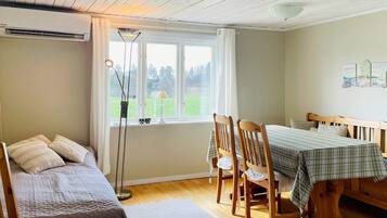 A room with a bed, a table with chairs, a window with a view of greenery, and an air conditioning unit.