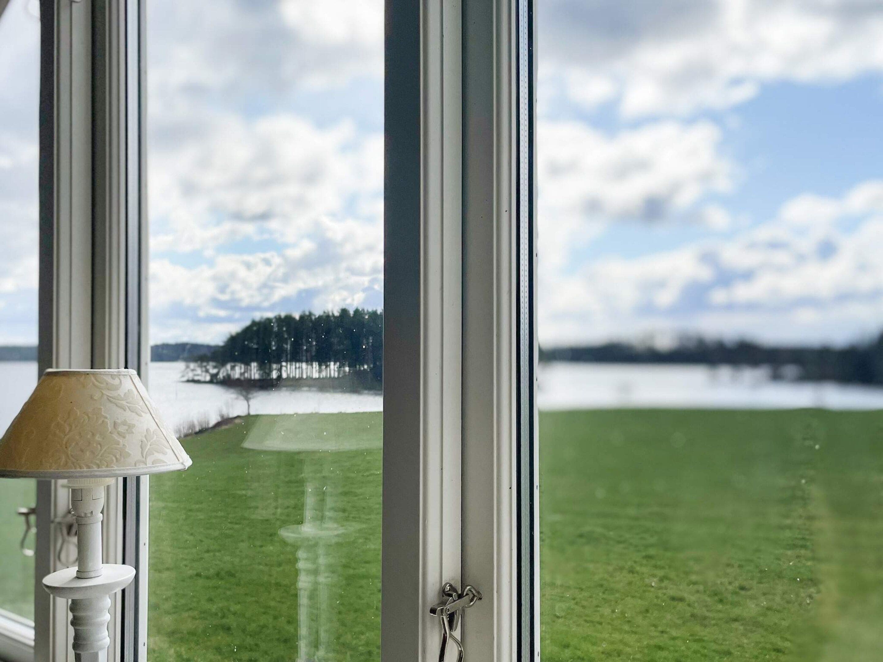 A view through a window with a lamp on the left, a door handle on the right, and a grassy landscape with trees and a body of water in the background.