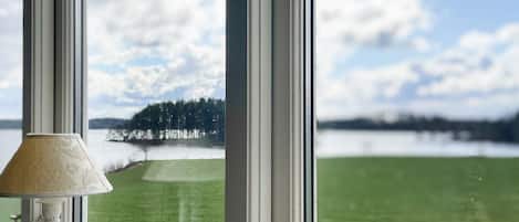 A view through a window with a lamp on the left, a door handle on the right, and a grassy landscape with trees and a body of water in the background.