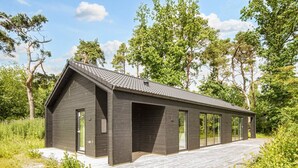 En moderne bygning i mørke farver med skråt tag og store vinduer, omgivet af vegetation og træer.