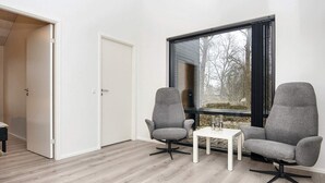 Et moderne værelse med to grå lænestole, et lille hvidt bord og et stort vindue med udsigt til træer.